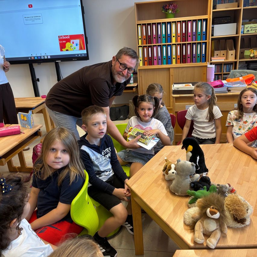 Eine Gruppe von Kindern sitzt um einen Tisch mit Kuscheltieren, während ein Mann ihnen in einem Klassenzimmer ein Buch vorliest.