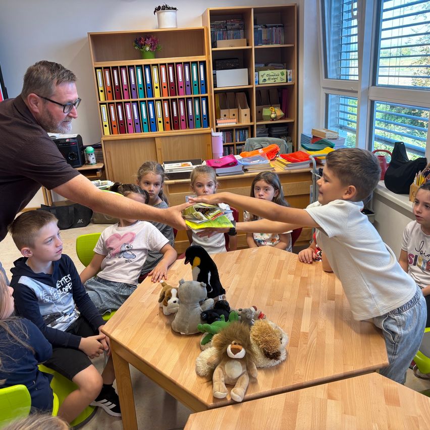 Eine Gruppe von Kindern sitzt um einen Tisch mit Kuscheltieren, ein Mann reicht einem Jungen ein Buch.