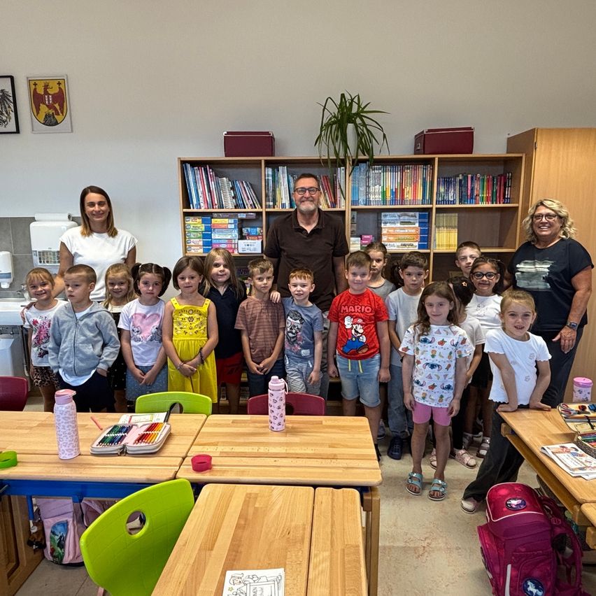 Eine Gruppe von Kindern und zwei Erwachsenen steht in einem Klassenzimmer mit Holztischen und Stühlen. Die Erwachsenen lächeln und stehen hinten. Die Kinder stehen vor den Tischen. Es gibt Bücher auf den Regalen und eine Topfpflanze in der Ecke.