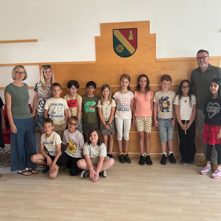 Eine Gruppe von Kindern und Erwachsenen steht zusammen in einem Raum. Sie tragen Freizeitkleidung. Zwei Frauen stehen an der Seite. Hinter ihnen ist eine Holzwand mit einem Logo.