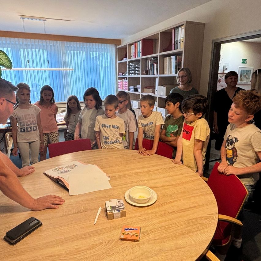 Eine Gruppe von Kindern und Erwachsenen versammelt sich um einen Tisch, möglicherweise in einem Klassenzimmer. Ein Mann steht, möglicherweise beim Unterrichten, während die Kinder zusehen. Der Tisch hat eine Schüssel, ein Buch und eine Schachtel.