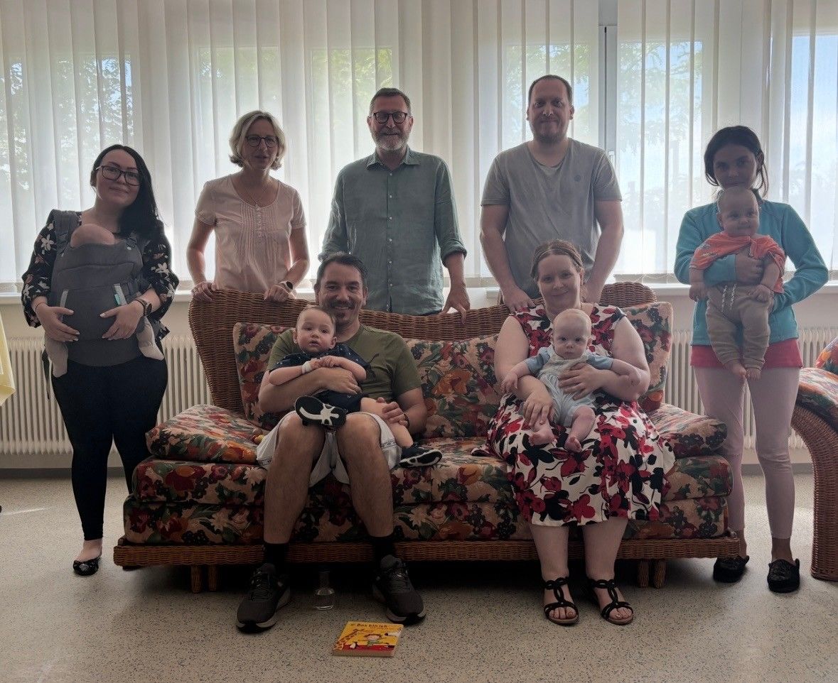 Eine Gruppe von Menschen posiert für ein Familienfoto in einem Raum mit Blumentapeten und weißen Jalousien. Zwei Erwachsene sitzen auf einem Sofa und halten Babys, während andere dahinter stehen.