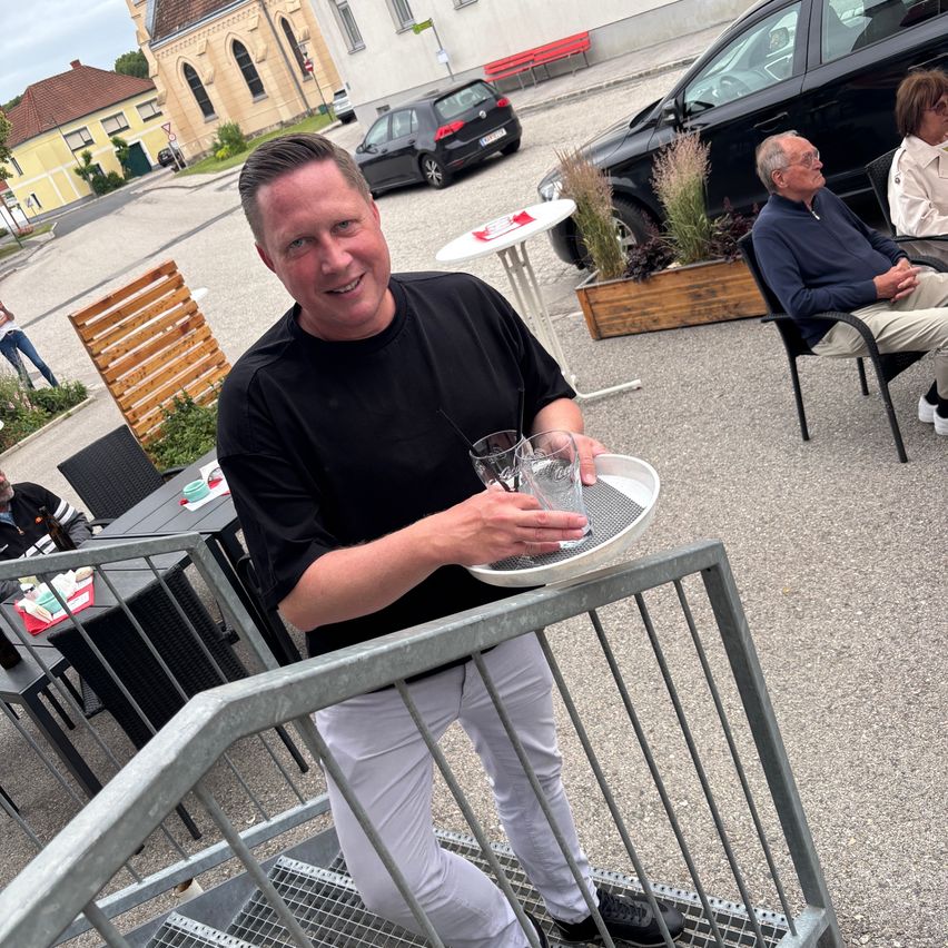 Ein Mann in einem schwarzen T-Shirt und weißen Hosen hält draußen vor einem Café einen Tablett mit Gläsern. Andere Gäste sitzen im Hintergrund, mit geparkten Autos dahinter.
