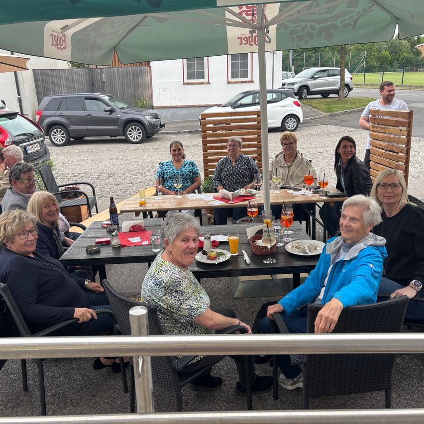 Eine Gruppe von Menschen sitzt um einen Tisch in einem Restaurant im Freien, mit Getränken und Tellern vor ihnen. Ein Regenschirm spendet Schatten.