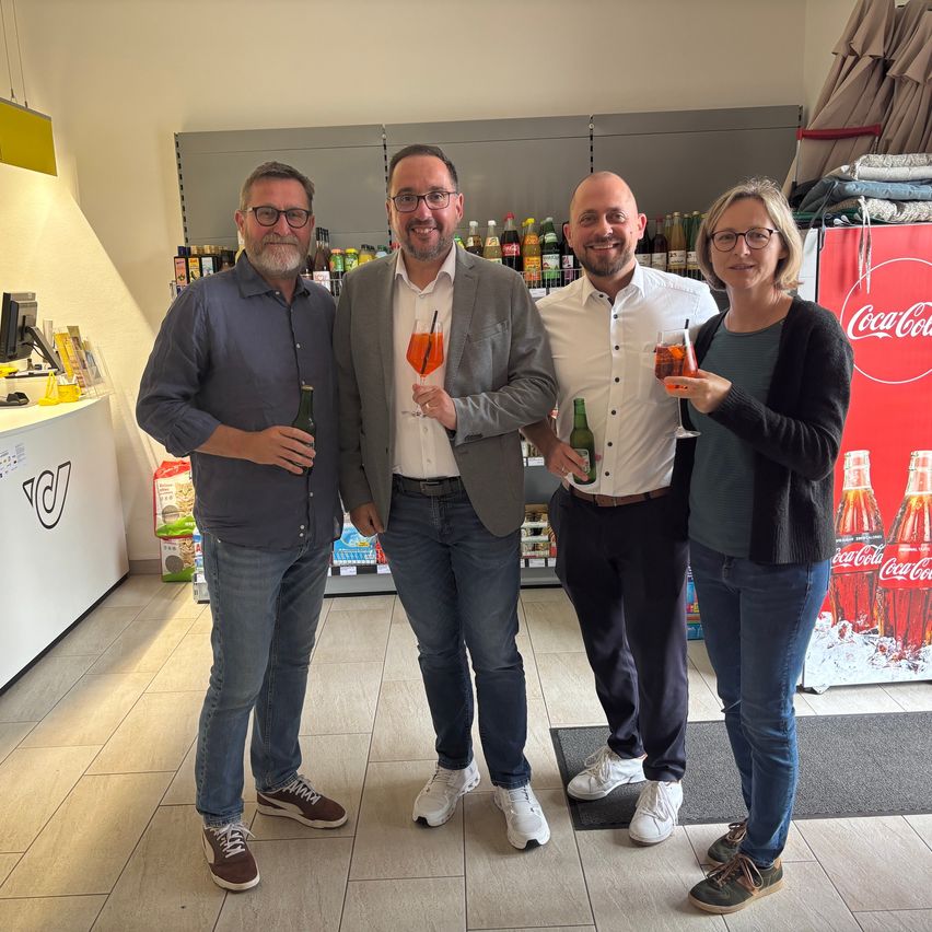 Vier Personen stehen lächelnd zusammen und halten Getränke. Ein Mann hält eine Flasche. Dahinter ist ein Regal mit vielen Flaschen. Rechts ist eine Coca-Cola Werbung.