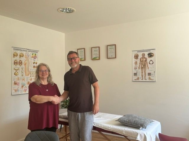 Ein erwachsener Mann und eine Frau schütteln sich in einem Raum mit medizinischen Diagrammen an der Wand die Hände.