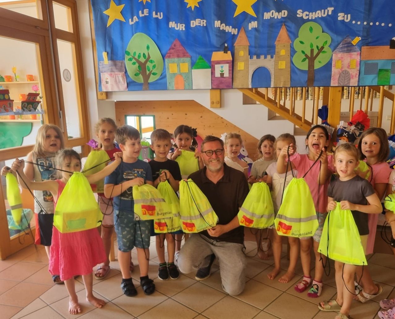 Eine Gruppe von Kindern und einem erwachsenen Mann in einem Klassenzimmer, die neonfarbene Taschen halten. Die Kinder lächeln und posieren für ein Foto. Hinter ihnen ist ein farbenfrohes Banner mit einer Zeichnung von Baum und Schloss.