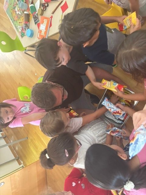Gruppe von Kindern und Erwachsenen sitzt im Kreis und teilt Snacks. Ein erwachsener Mann mit Brille hält einen Behälter mit Snacks.