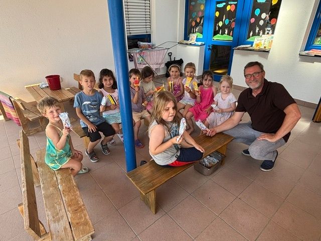 Eine Gruppe von Kindern und ein erwachsener Mann sitzen auf Bänken und halten etwas in den Händen. Dahinter steht ein Tisch mit einem Eimer und Büchern darauf.