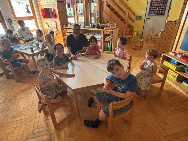 Eine Gruppe von Kindern sitzt um einen Tisch mit einem Mann in einem Raum. Sie lächeln und scheinen für ein Foto zu posieren.
