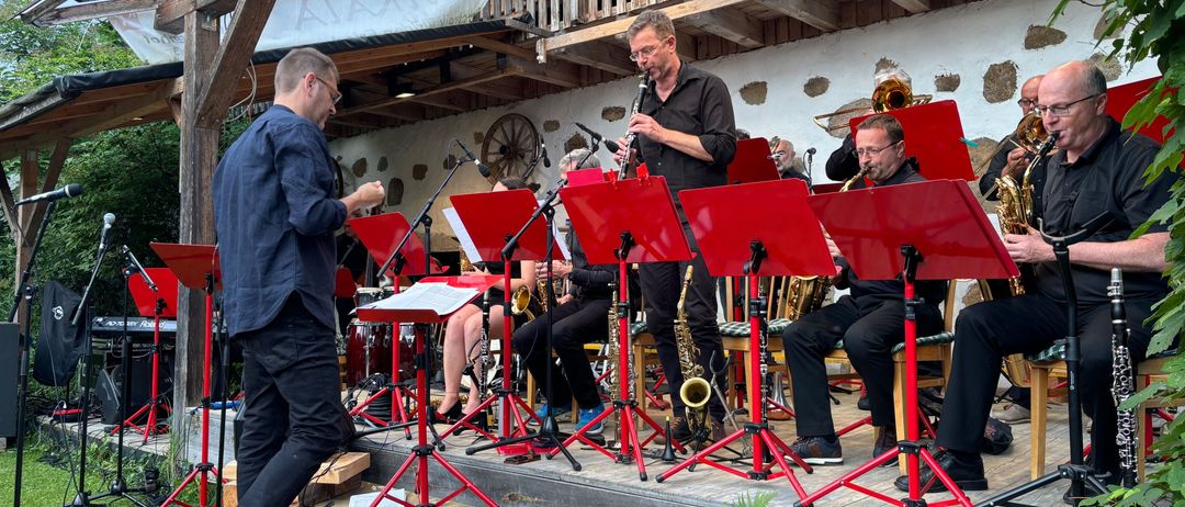 Eine Band proben auf einer hölzernen Bühne unter einem Dach. Ein Dirigent führt von vorne, während Musiker verschiedene Instrumente spielen. Dahinter ist ein hölzerner Balkon sichtbar.