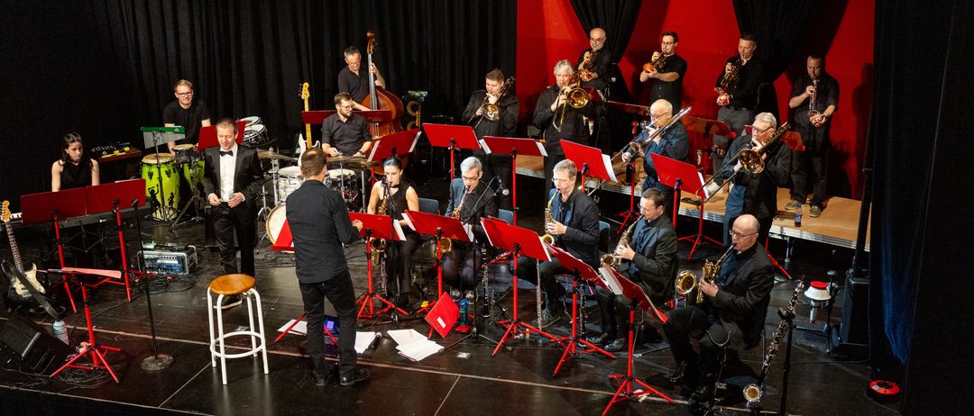 A jazz band performs on stage, with musicians playing trumpets, saxophones, and other instruments. A man stands and directs, while others sit and play in rows.