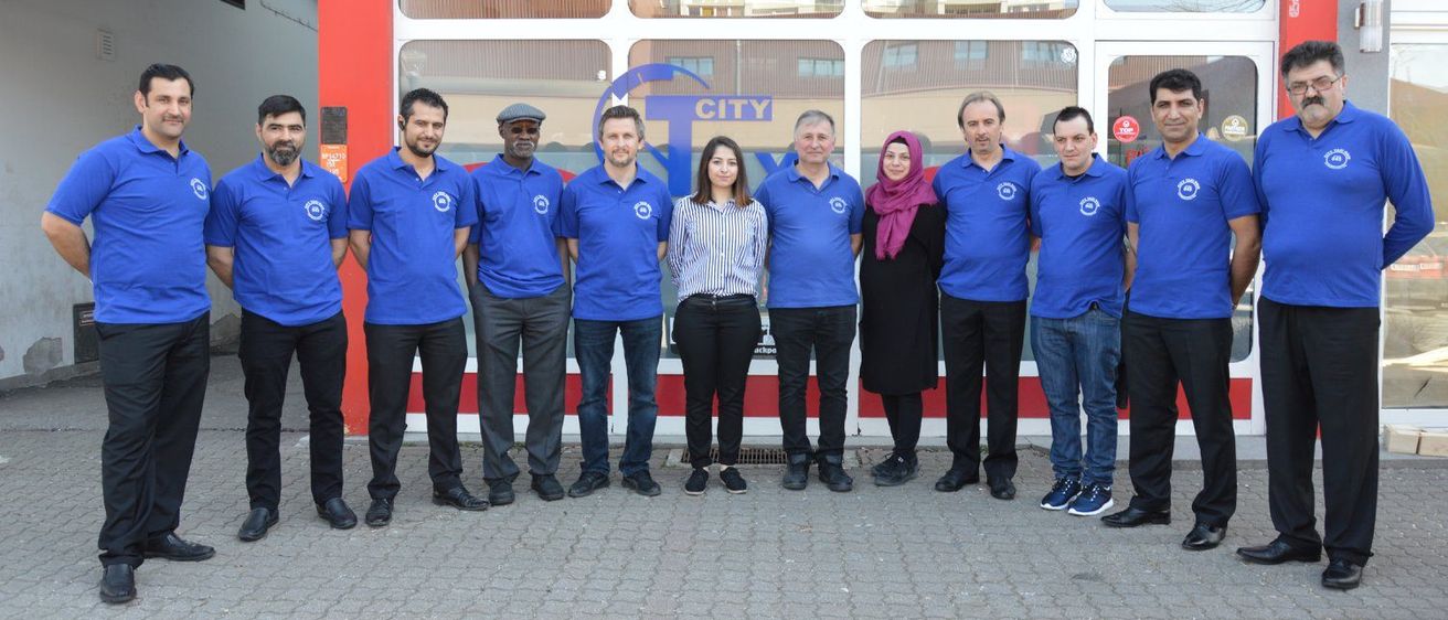 Eine Gruppe von Menschen in blauen Hemden und schwarzen Hosen steht vor der Fassade eines Taxiunternehmens.