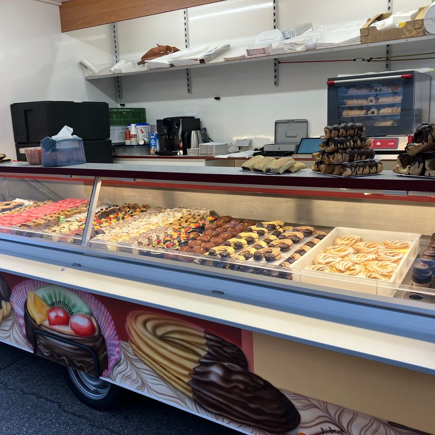 Eine Vitrine mit verschiedenen Desserts wie Donuts, Gebäck und mehr, in einer Bäckerei. Hinter der Vitrine befinden sich Regale mit Zutaten.