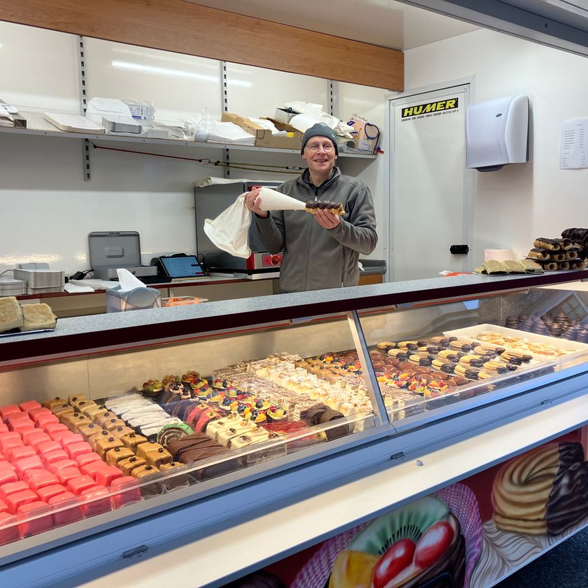 Ein Mann steht hinter einer Bäckereitheke und hält eine Gebäcktüte. Die Theke ist mit verschiedenen Desserts gefüllt. Über ihm befinden sich Regale mit Gegenständen und eine Tür mit der Aufschrift 'HUMER'.