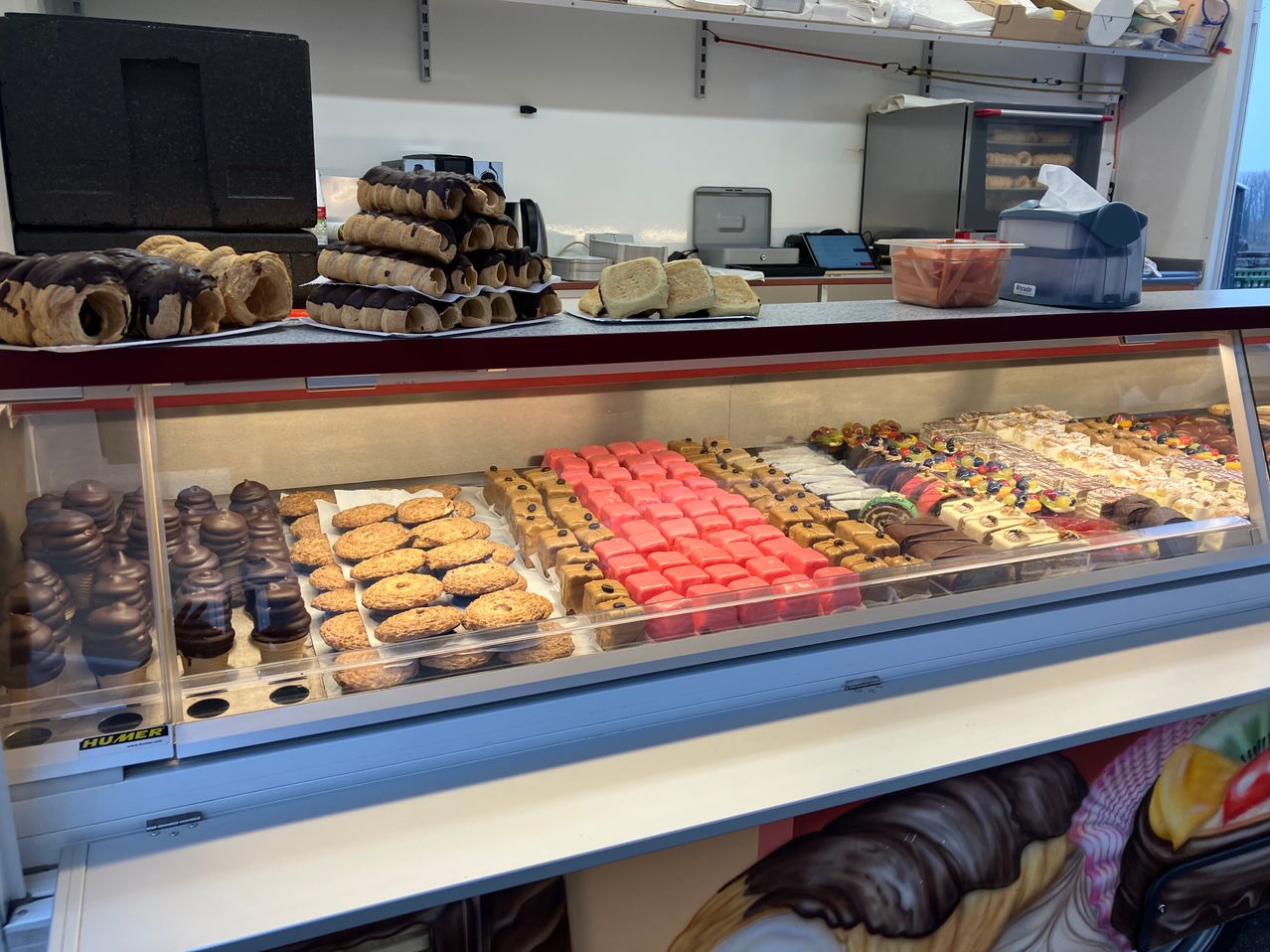 Eine Bäckerei-Vitrine mit verschiedenen Gebäck und Desserts. Oben gibt es Stapel von schokoladenüberzogenen Gebäckstücken und Kuchenscheiben. Unten sind Reihen von Keksen, Kuchen und anderen süßen Leckereien.