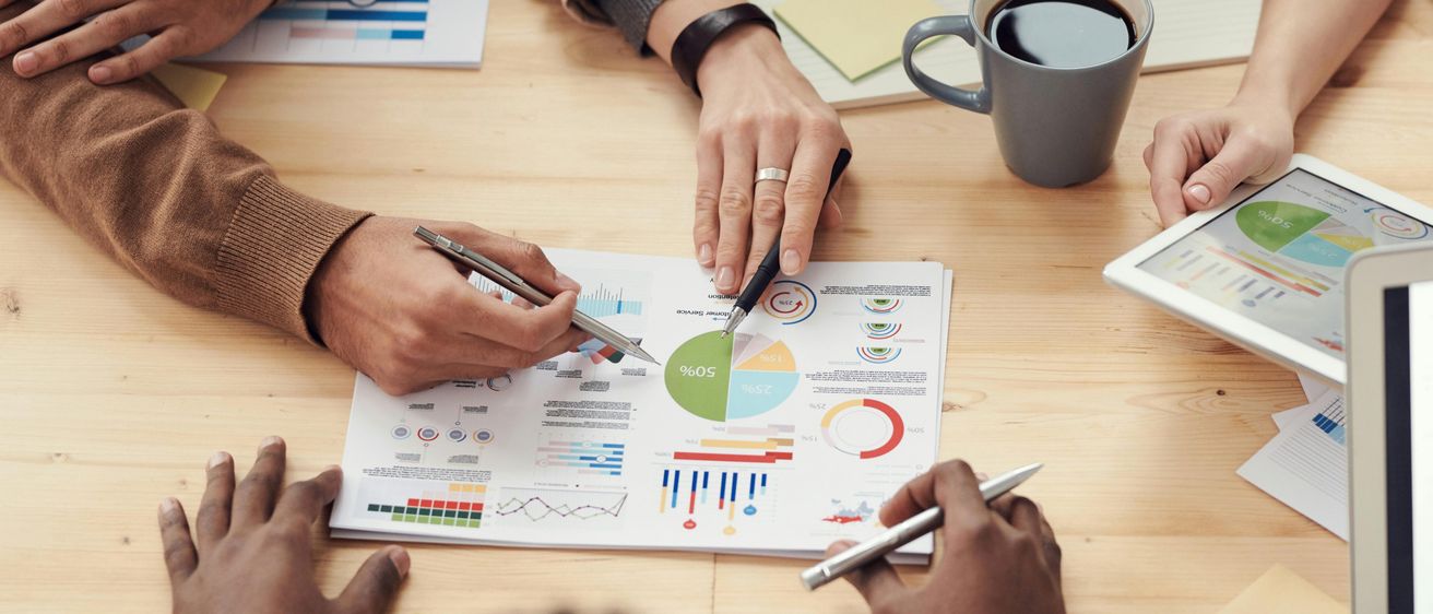 Zwei Personen betrachten ein Papier mit Grafiken auf einem Holztisch. Eine Person hält einen Stift und schreibt. Ein Kaffeebecher steht auf dem Tisch.