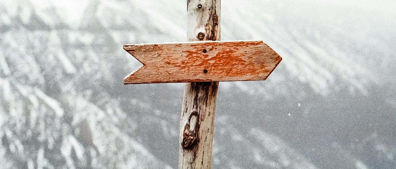 Ein hölzernes Wegweiser mit einem nach rechts zeigenden Pfeil steht vor einem verschwommenen Hintergrund mit schneebedeckten Bergen.