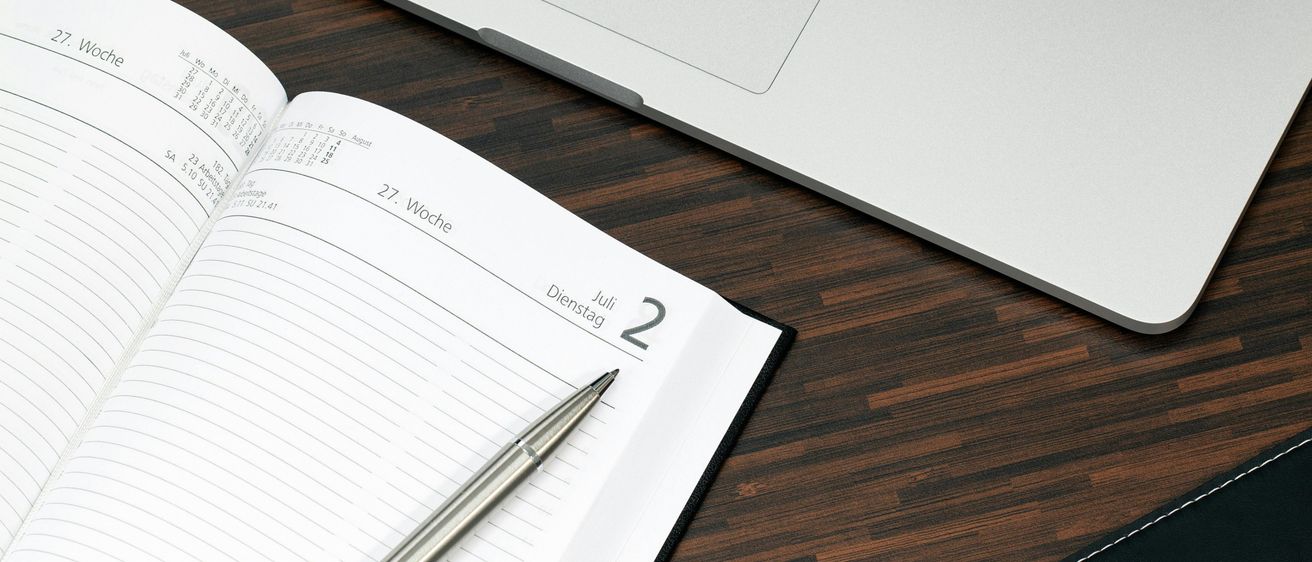 Ein offenes Kalender auf einem hölzernen Schreibtisch mit einem Stift darauf. Ein Laptop ist hinter dem Kalender platziert. Der Kalender zeigt die 27. Woche und den 2. Juli, der ein Dienstag ist.