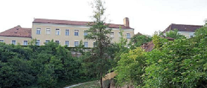 Ein kleiner Kiesweg führt durch einen grasbewachsenen Bereich mit dichten Sträuchern und Bäumen. Hinter der Begrünung befindet sich ein großes Gebäude mit mehreren Fenstern und einem roten Dach.