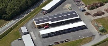 Luftaufnahme einer Anlage mit Solarpanelen auf den Dächern, umgeben von Bäumen und einer Straße, mit mehreren Fahrzeugen auf dem Parkplatz.