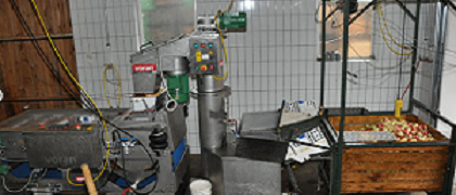 Eine Industrieküche mit einer Metallmaschine und einem hölzernen Apfelkorb. Die Maschine hat mehrere Knöpfe und Kabel. Der Raum hat geflieste Wände und eine Holztür.