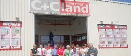 Eine Gruppe von Menschen steht vor einem Laden. Der Laden hat eine weiße Wand und ein rotes Schild, das C+C thermen land steht.