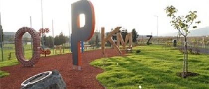 Ein Park mit einem roten Weg, der einen großen Buchstaben 'P', eine Reifenskulptur und verschiedene Spielgeräte zeigt. Ein Baum und ein großes 'O' sind links. Der Himmel ist klar, und der Bereich ist von einem Zaun umgeben.