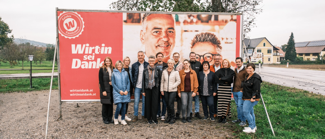 Eine Gruppe von Menschen steht vor einem großen roten Schild mit den Worten 'Wirtin sei Dank' und einem Bild eines lächelnden Mannes. Sie alle lächeln und scheinen für ein Foto zu posieren.