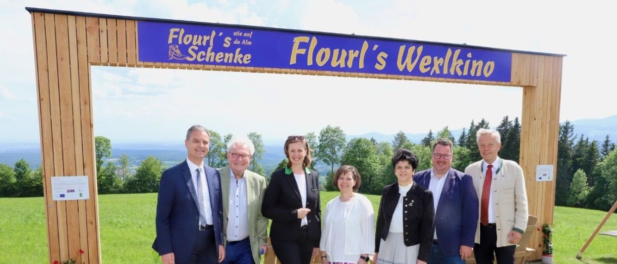 Eine Gruppe von Menschen steht unter einem Schild, das 'Flourl's Werxlino' besagt. Sie sind in Geschäftskleidung und scheinen für ein Foto zu posieren. Hinter ihnen ist ein Rasenfeld mit Bäumen und Blick auf die Berge.