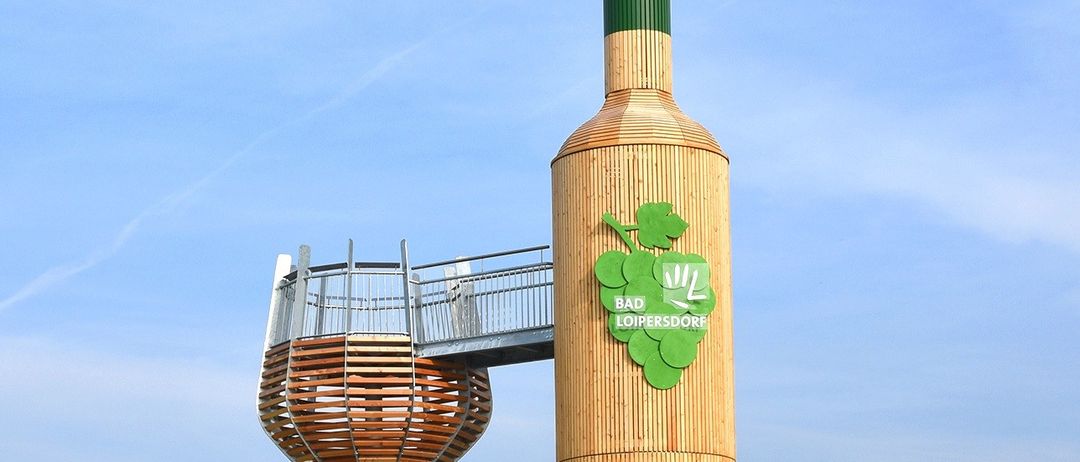 Eine große hölzerne Flaschenskulptur steht auf einem grünen Hügel mit einer Terrasse im Inneren der Flasche. Die Flasche hat einen grünen Deckel und ein Etikett, das Bad Loipersdorf liest.