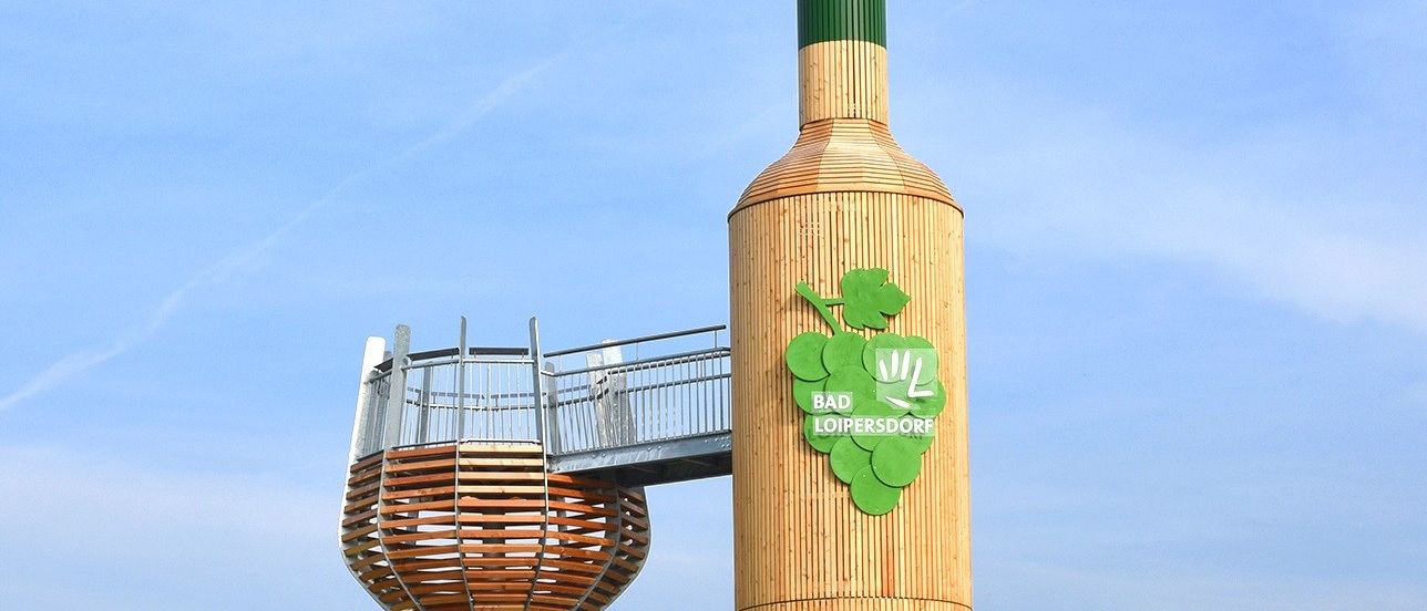 Eine große hölzerne Flaschenskulptur steht auf einem grünen Hügel mit einer Terrasse im Inneren der Flasche. Die Flasche hat einen grünen Deckel und ein Etikett, das Bad Loipersdorf liest.