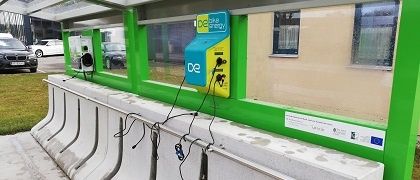 Eine grüne und weiße Bushaltestelle mit einem blau-gelben Zahlungsautomaten, der mit 'DE' beschriftet ist. Es gibt mehrere Ladestationen für Elektrofahrzeuge. Im Hintergrund sind geparkte Autos und ein Gebäude zu sehen.
