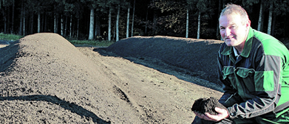 Ein Mann in einer grünen und schwarzen Jacke kniet auf einem Erdfeld und hält ein Stück Erde in den Händen. Bäume und Pflanzen sind im Hintergrund. Das Foto wurde von Peter Ringhofer-Recherberger aufgenommen.