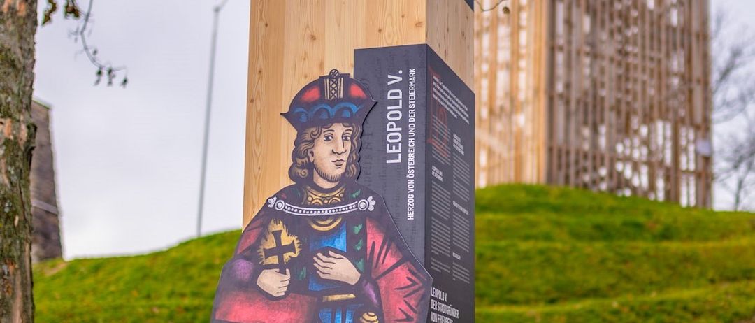 Ein hölzerner Pfosten mit einer Wandmalerei eines Mannes, vermutlich Leopold V., steht auf einem Rasenhügel. Dahinter ist ein Turm zu sehen.
