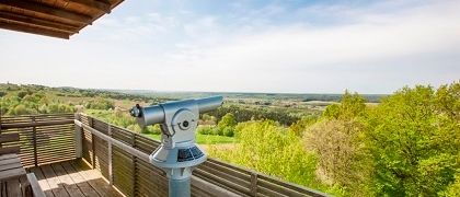 Ein Teleskop ist auf einer Holzterrasse montiert und bietet einen Panoramablick auf ein grünes Tal unter einem klaren blauen Himmel mit flauschigen Wolken.