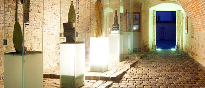 Ein schwach beleuchteter Ziegeldurchgang mit einer Reihe von Glasvitrinen auf der linken Seite. Jede Vitrine enthält Skulpturen und Artefakte unter heller Beleuchtung. Die rechte Seite des Tunnels führt zu einer offenen Tür, die von einem gelben Licht beleuchtet wird.
