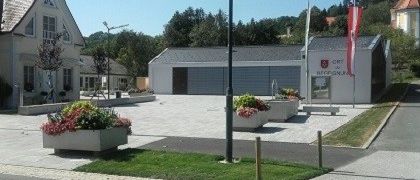 Ein Gebäude mit Garage und Flaggen steht vor einer Straße. Blumentöpfe und Blumen sind um den Eingang herum angeordnet. Ein Haus und ein weiteres Gebäude sind im Hintergrund zu sehen.