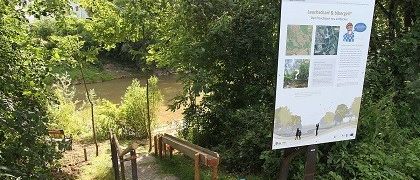 Ein Schild steht neben einer Holzbank in der Nähe eines Flusses mit Stufen, die zu ihm hinaufführen. Es gibt zwei blaue Eimer auf dem Boden. Bäume und Pflanzen umgeben das Gebiet.