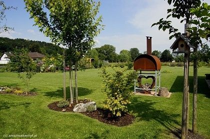 Ein Garten mit einem herzförmigen Pizzaofen, umgeben von Bäumen, Büschen und Steinen. Ein Haus und Bäume sind in der Ferne.