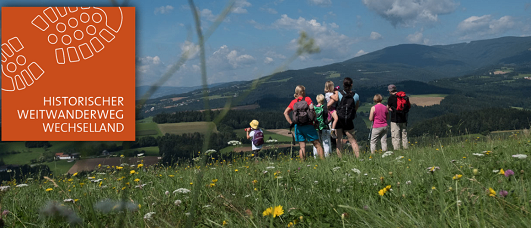 Eine Gruppe von Wanderern steht auf einem Grashügel und blickt auf eine Bergkette. Sie sind mit Rucksäcken und Hüten ausgestattet. Das Bild hat einen Wasserzeichen in der oberen linken Ecke mit dem Text 'Historischer Weitwanderweg Wechselland'.