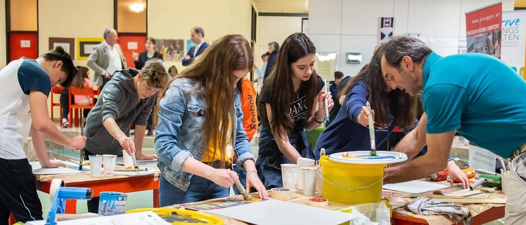 Eine Gruppe junger Menschen arbeitet in einem Klassenzimmer an einem Kunstprojekt und verwendet dabei Pinsel und Eimer mit Farbe. Einige Personen beobachten im Hintergrund.