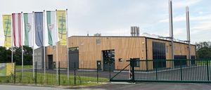 Ein modernes, großes Gebäude mit Holzfassade und Solarpanelen ist zu sehen. Fahnen sind draußen gehisst und ein Tor ist offen. Ein Parkplatz ist davor mit einem Rasenbereich.