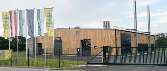 Ein modernes, großes Gebäude mit Holzfassade und Solarpanelen ist zu sehen. Fahnen sind draußen gehisst und ein Tor ist offen. Ein Parkplatz ist davor mit einem Rasenbereich.