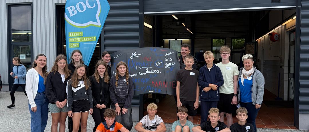Eine Gruppe junger Menschen posiert für ein Foto vor einem Gebäude mit den Worten 'BOC' und 'Service' darauf. Sie stehen und sitzen auf dem Boden, lächeln und schauen in die Kamera. Hinter ihnen steht eine Tafel mit Zeichnungen.