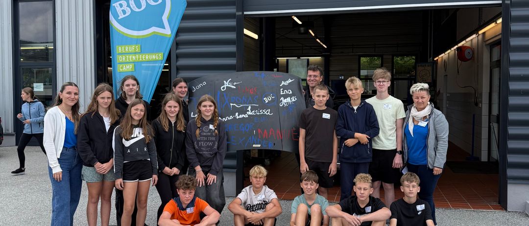 Eine Gruppe junger Menschen posiert für ein Foto vor einem Gebäude mit den Worten 'BOC' und 'Service' darauf. Sie stehen und sitzen auf dem Boden, lächeln und schauen in die Kamera. Hinter ihnen steht eine Tafel mit Zeichnungen.