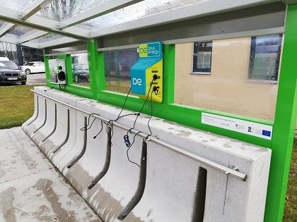 Eine grüne Bushaltestelle mit einer DE-Ladestation an der Wand, gekennzeichnet durch ein gelbes und blaues Schild. Eine Betonbank ist an der Haltestelle befestigt, und ein Auto ist in der Nähe geparkt.