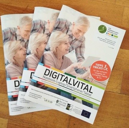 Drei Broschüren mit dem Titel 'DigitalVital' liegen auf einer Holzoberfläche. Sie zeigen Bilder älterer Erwachsener und präsentieren einen Laptop, ein Smartphone und ein Tablet. Die Broschüren bieten Tipps und Tricks zur digitalen Kompetenz.