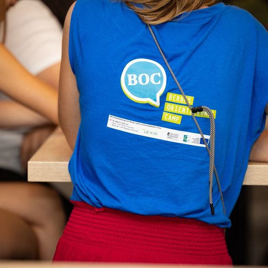 Eine Frau trägt ein blaues Shirt mit den Buchstaben BOC und einer Sprechblase. Ein weißer Aufkleber befindet sich auf dem Rücken.