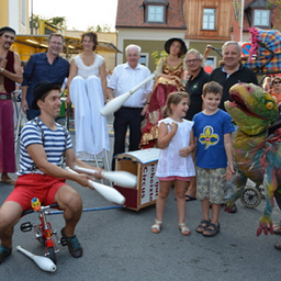 Eine Gruppe von Menschen steht auf einer Straße. Ein Mann jongliert mit Stöcken, und ein Mädchen in einem weißen Shirt steht neben ihm. Hinter ihnen sind ein Dinosaurierkostüm und ein Kinderwagen zu sehen. Menschen stehen hinter ihnen, und Gebäude sind im Hintergrund.